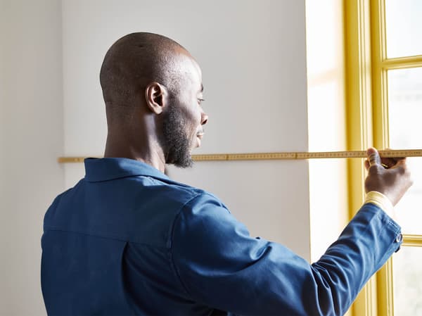 A man with a blue jacket is measuring a wall next to the window.