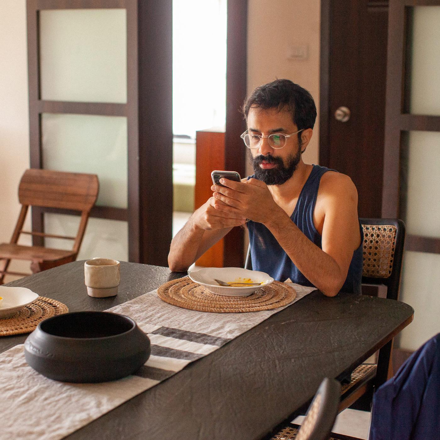 A man with dark hair and a beard just finished eating and is browsing his mobile phone at the kitchen table.