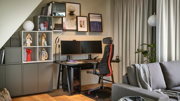 A MATCHSPEL gaming chair and UTESPELARE gaming desk in the corner of a grey room holding two monitors and other accessories.