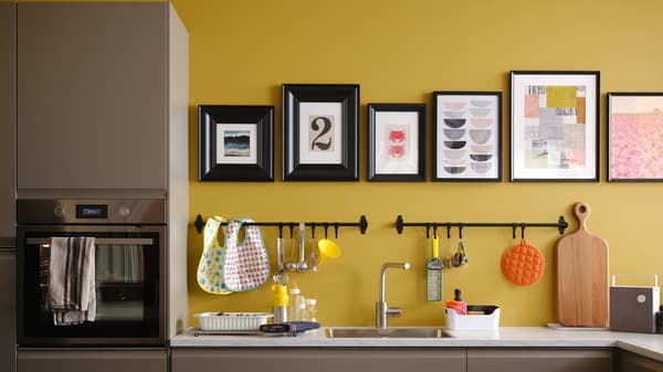 A matt dark beige kitchen with a white marble effect worktop, plus black rails and pictures in black frames on a yellow wall.