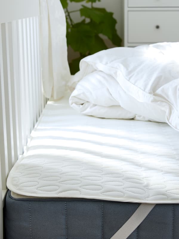A mattress, with a mattress protector on top of it, lies on a bed frame. A crumpled duvet lies on the mattress protector.