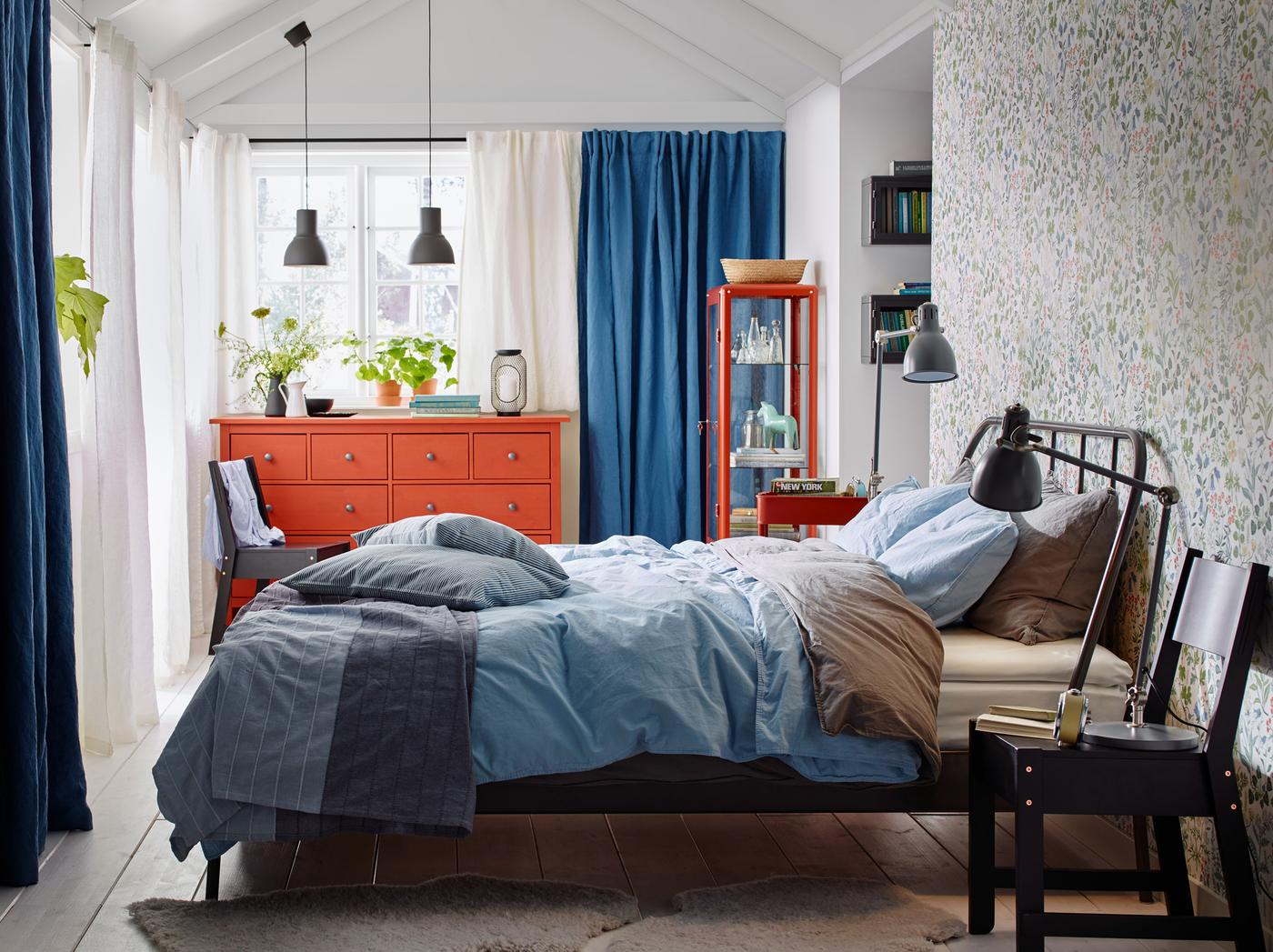A medium sized bedroom with a dark grey steel bed for two, with bedlinen in light blue and grey. Shown together with a red chest of drawers with four small drawers and four large drawers.