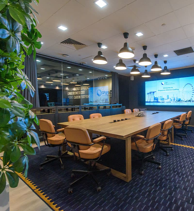 A meeting room setting featuring two long wooden tables and ten light brown swivel chairs on two dark blue rugs and a plant wall at the the back of the room.