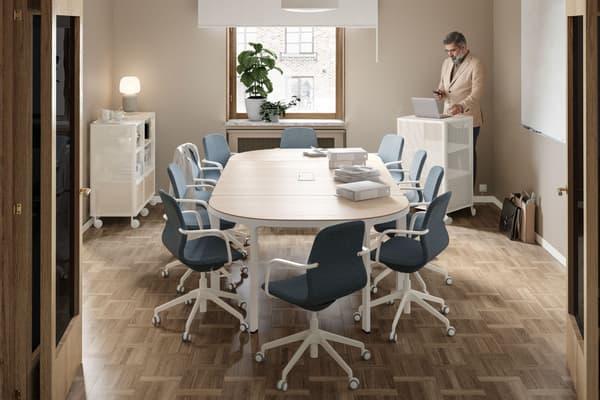 A meeting room with a conference table and 10 chairs. A man stands at the back by a window, behind a computer on a cabinet.