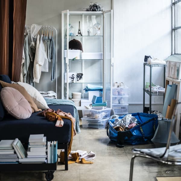 A messy room with sofa, white shelving unit, curtain, stacked books, an IKEA blue bag of stuff on the floor and window light.