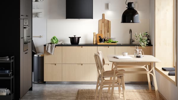 A METOD kitchen with FRÖJERED kitchen fronts in light bamboo, a table and two chairs and a SVARTNORA pendant lamp in black.