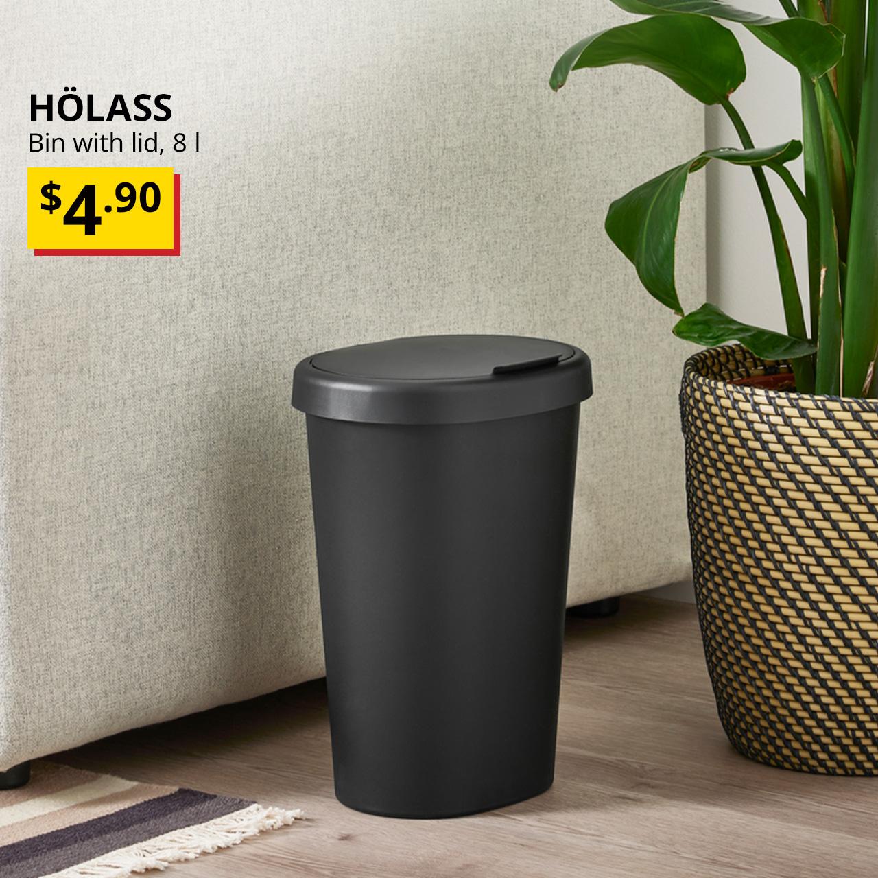 A mid shot of a black HÖLASS bin with lid next to a sofa and a plant pot in the living room setting. On the top left corner, black text overlay image that reads "HÖLASS, Bin with lid, 8 l", followed by a breath-taking price tag in yellow/red with price of $4.90 in black.