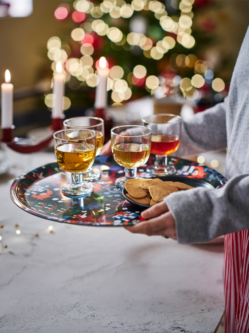 A mid shot of a lady holding a multicolour VINTERFINT tray with a handful of STOCKHOLM 2025 wine glasses in the dining room.