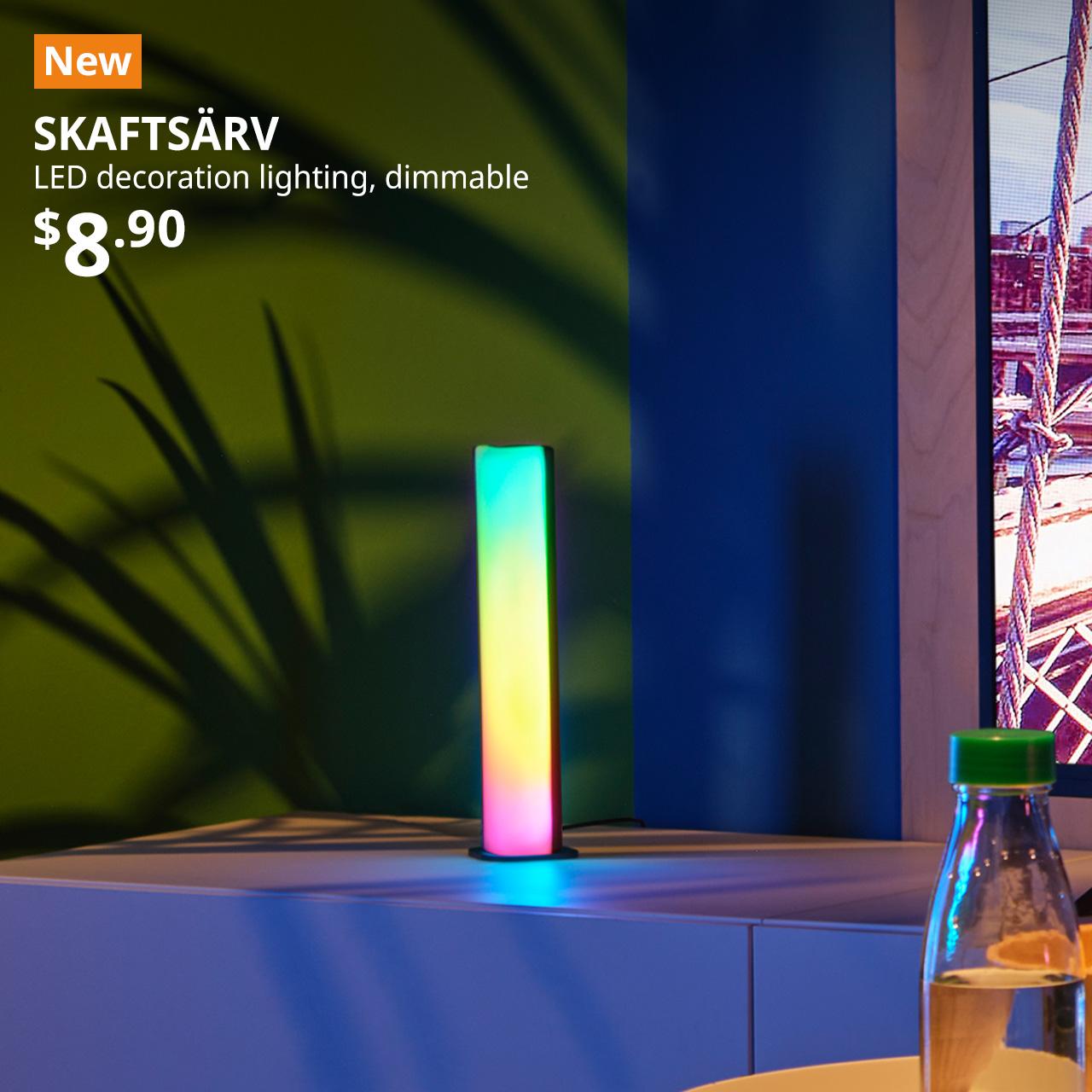 A mid shot of a SKAFTSÄRV LED decoration lighting placed on a white BESTÅ TV console next to a TV in the living room setting. On the top left corner, an orange tag with white text that reads 'New', followed by white text overlay image that reads 'SKAFTSÄRV, LED decoration lighting, dimmable, $8.90'.