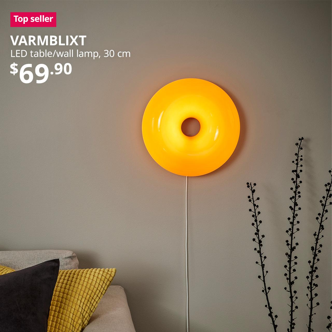 A mid-shot of an orange VARMBLIXT LED table/wall lamp being hung on the wall next to a faringe light grey FRIHETEN sofa in the living room setting. A pink/red tag with white text that reads "Top seller", followed by white text overlay image that reads, "VARMBLIXT, LED table/wall lamp, 30 cm, $69.90"
