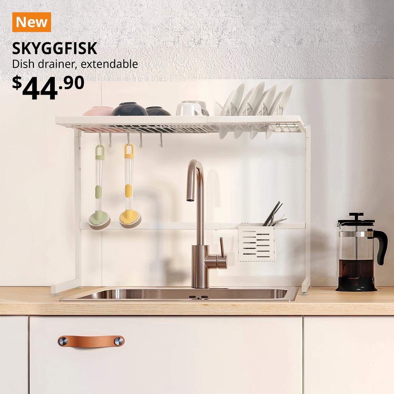 A mid shot of the kitchen counter's sink area where it feature a white SKYGGFISK dish drainer being utilised over the sink. An orange "NEW" label is displayed in the top left corner of the image, with bl;ack text overlaying image that reads 'SKYGGFISK, Dish drainer, extendable, $44.90'.