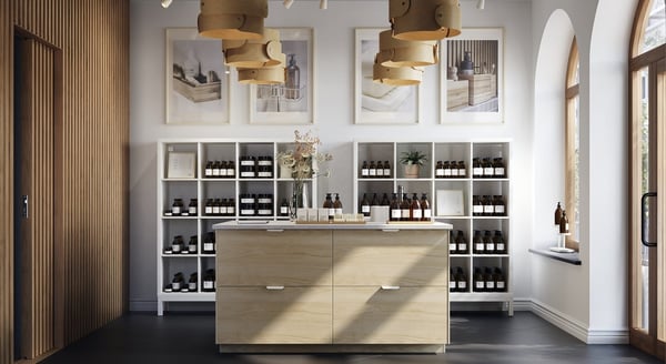 A minimalist and elegant boutique interior featuring natural wood and white tones. At the center is a wooden island with six drawers and a white countertop, displaying amber glass bottles, soap bars, and a vase of dried flowers. Behind the island, two symmetrical white shelving units house rows of neatly arranged brown bottles and a few decorative items. Above the shelves hang five framed prints showcasing beauty products and textures. The space is well-lit with modern track lighting and unique circular wooden pendant lamps. Large arched windows with wooden frames allow natural light to flood the shop, adding warmth and serenity.