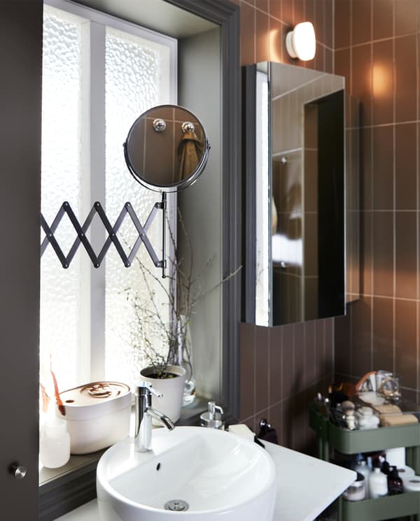 A mirror with an extension arm that’s pulled out over a sink cabinet, a cabinet with mirror door and a gray-green trolley.