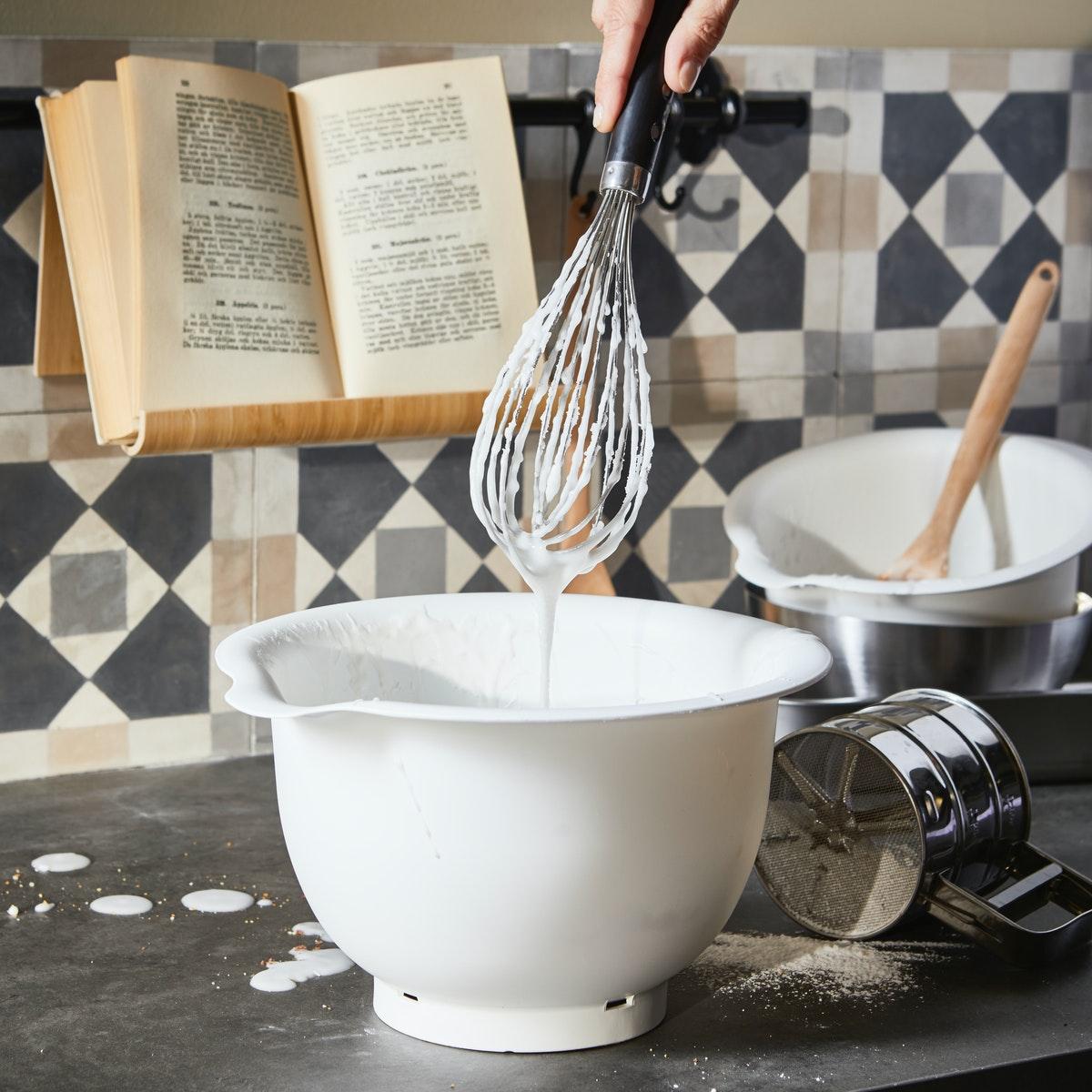 A mixing  bowl positioned on a messy counter  top. Ready to start baking. A recipe book can be seen in the back ground.