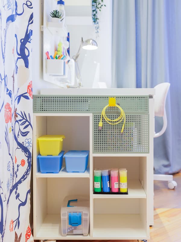 A mobile OVNING trolley with open shelves, colourful storage boxes and hanging accessories, placed beside a desk in a bright and tidy study space.