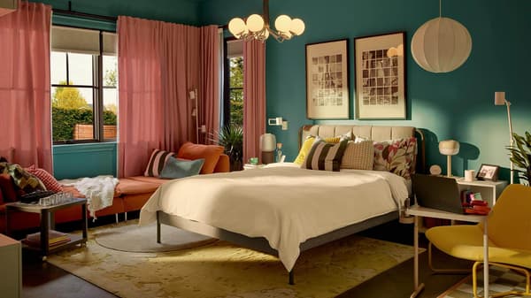 A modern bedroom with a bed, colorful pillows, and artwork. An orange sofa with books to the left, a yellow armchair to the right, and a white floor lamp near the window.