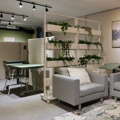 A modern co-working space with a lounge area featuring a large grey sofa, armchairs, soft lighting, and indoor plants. Open workstations, neutral tones, and industrial-style details create a calm and welcoming environment for work, meetings, and relaxation.