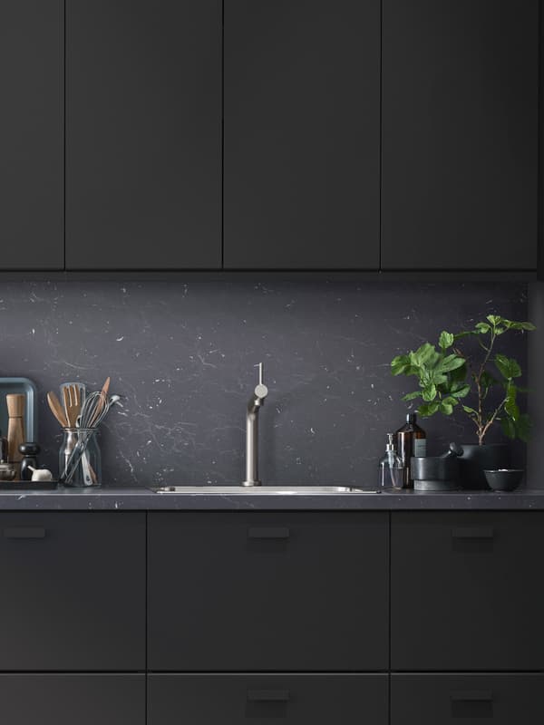 A modern kitchen with black KUNGSBACKA fronts and utensils on one side of the sink and a large plant on the other.