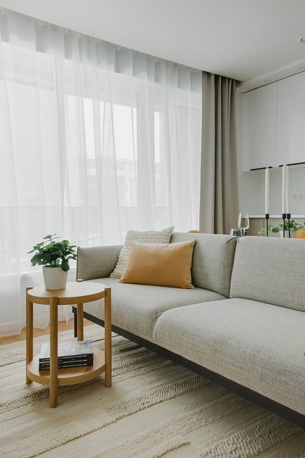 A modern living room corner featuring a light gray fabric sofa with a mustard yellow pillow and a textured cream cushion. Next to the sofa is a small round wooden side table holding a potted plant and a few stacked books. Soft, sheer white curtains allow natural light to fill the space, while beige blackout curtains frame the window. The neutral-toned textured rug beneath enhances the cozy and minimalist aesthetic.