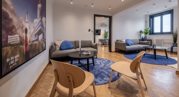 A modern lounge space with parquet flooring, blue round rugs, two gray sofas, light wooden chairs, and black round coffee tables. The room features neutral walls, plants near the windows, and a large painting on the left wall showing a boy watching a rocket launch with Lithuanian text. A doorway in the center leads to a meeting area with more seating.