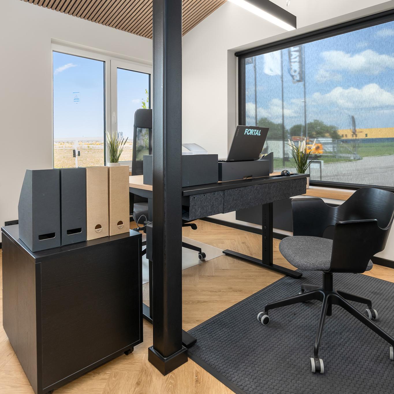 A modern office interior featuring a sleek sit/stand desk setup with a laptop, office chairs, and neatly organised magazine files on a cabinet beside large windows letting in natural light.