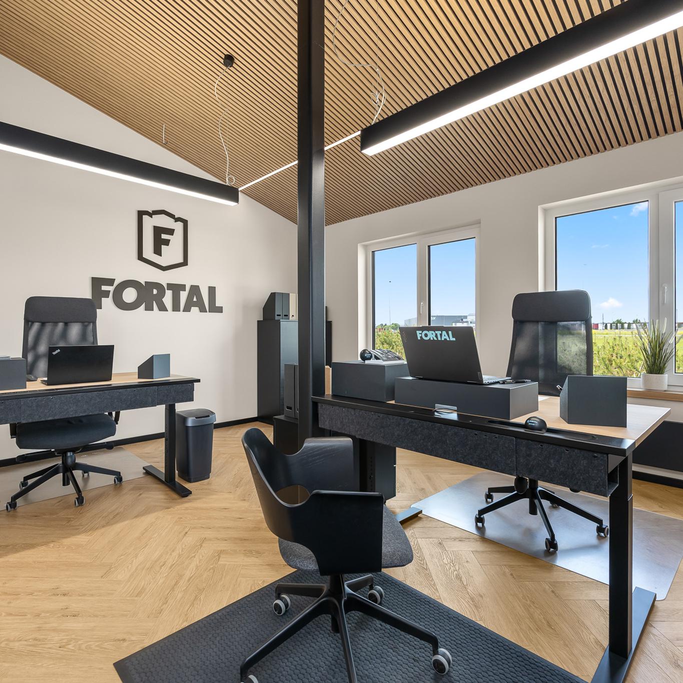 A modern office workspace with two sleek sit/stand desks, ergonomic chairs, and neatly organised office accessories is arranged on light wood flooring under minimalist linear ceiling lights. Large windows bring in natural light and reveal a view of a grassy industrial area.