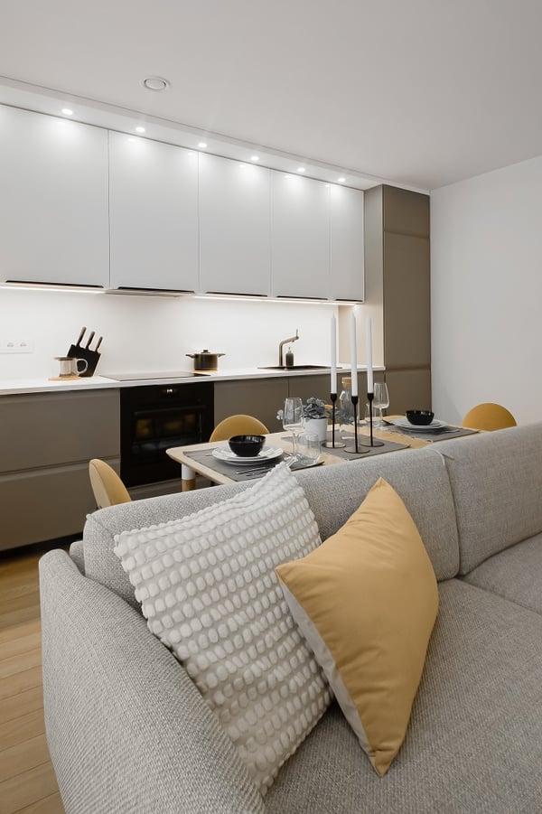 A modern open-plan kitchen and dining area featuring sleek white upper cabinets with under-cabinet lighting and matte taupe lower cabinets. A minimalist dining table is set with black bowls, glassware, and tall white candles. The foreground includes a light gray sofa adorned with textured white and mustard-yellow pillows, adding warmth and contrast to the clean, neutral interior.