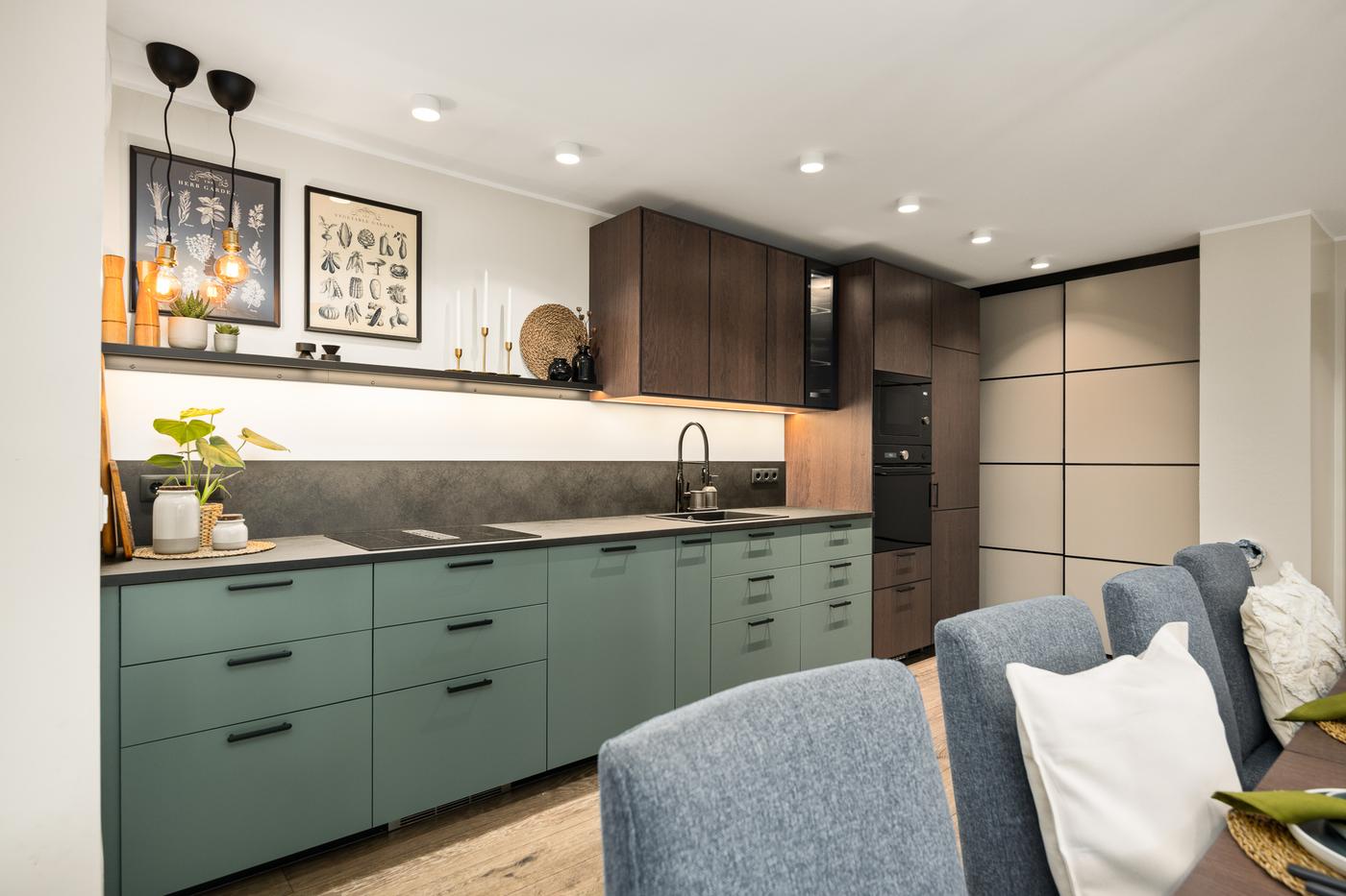 A modern open-plan kitchen with darker cabinets and a light worktop. The lower cabinets are in a green-grey tone. The space is clean, well-organised, and blends into the living area.