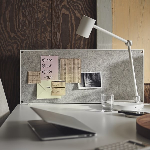 A modern white desk set against a dark wood paneled wall features a minimalist workspace with a soft gray felt pinboard. Pinned to the board are various notes: a pink sheet with handwritten calculations, a vintage data chart, a Sudoku puzzle, a yellow envelope with a note, a waveform graph, and a black-and-white photo of an architectural corridor. A sleek white desk lamp with an adjustable arm illuminates the area. On the desk lie a partially closed laptop, a clear drinking glass, a wooden tray, and a keyboard, creating a clean and functional office environment.