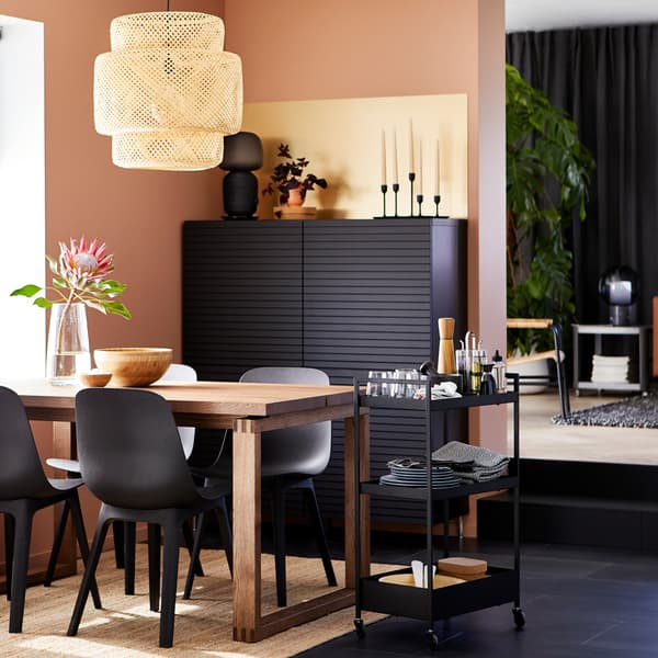A MÖRBYLÅNGA oak veneer table with black ODGER chairs by a window, with a black cabinet and a trolley holding tableware.