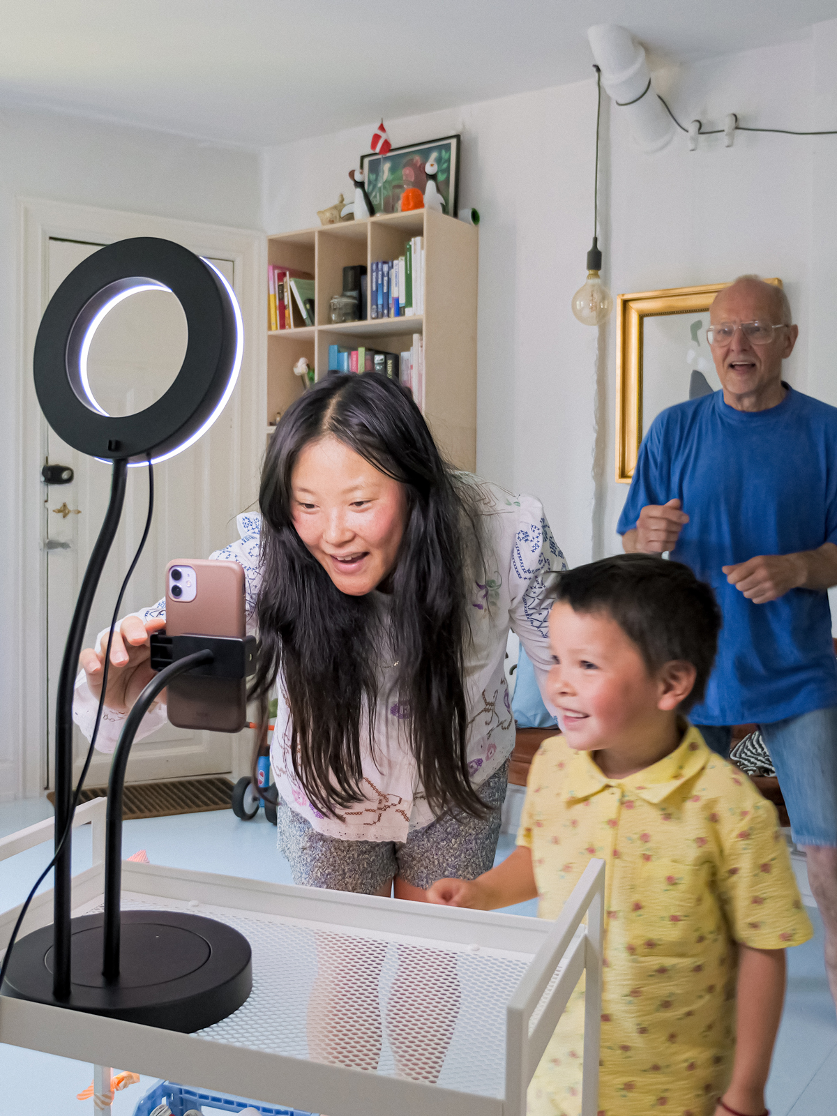 A mother and her child set up a LÅNESPELARE ring light with phone holder, ready to record a video. The father dances in the background.