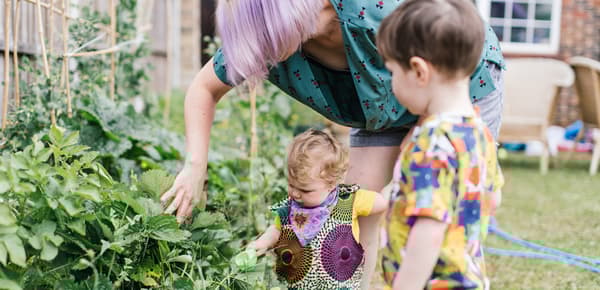 A mother and her two children are standing in their backyard. She is looking at a plant and moving its leaves with her hand.