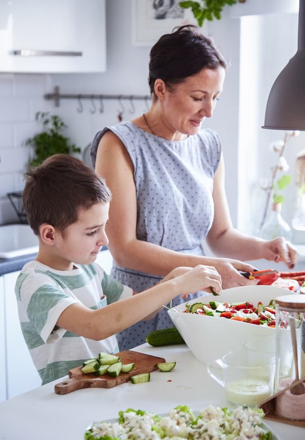 A mother and her young son prepare a salad together in a white kitchen.