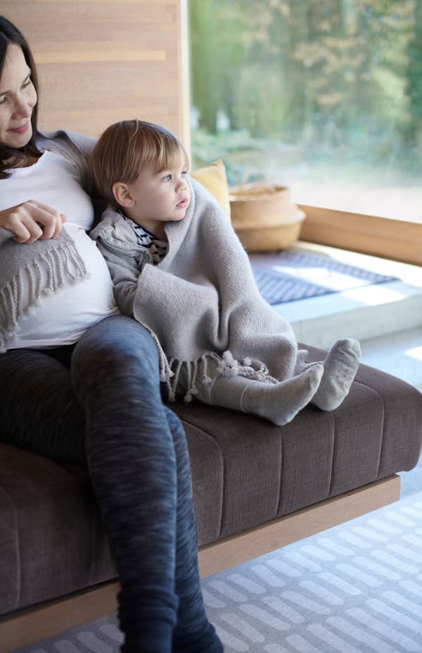 A mother and young child wrapped in a grey OMTÄNKSAM wool throw sit close together on a sofa.