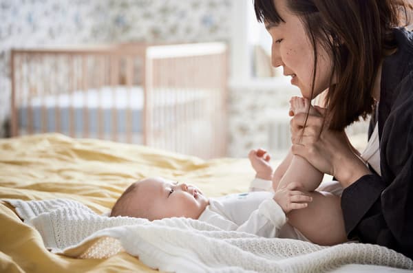 A mother holds her baby's toes and is smiling as the baby lies on yellow bedsheets with a crib in the background.