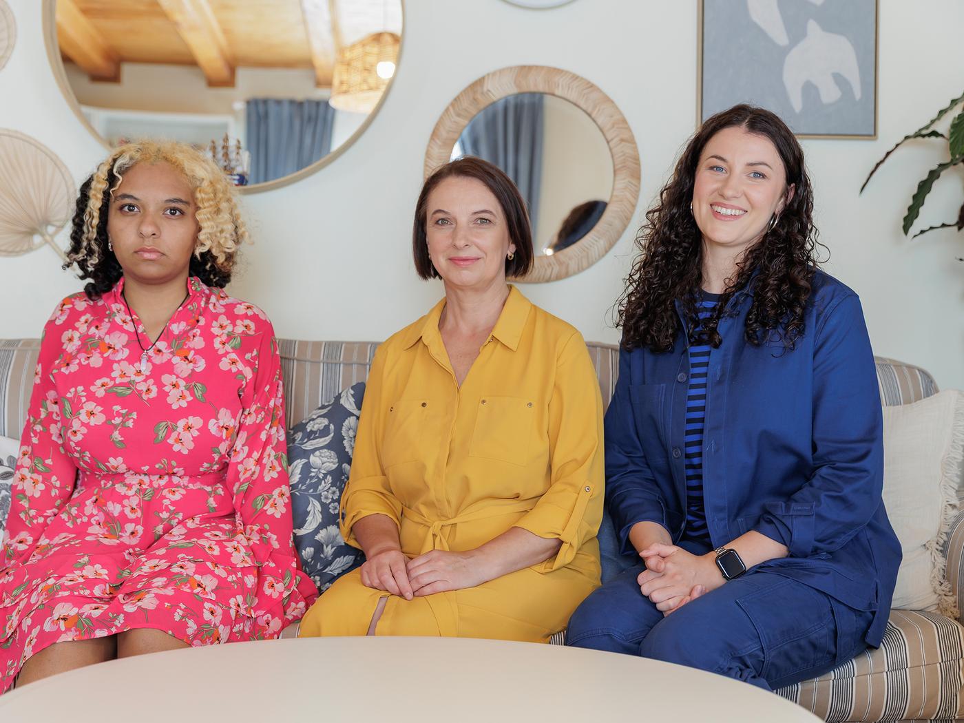 A mother in a yellow dress, daughter in a red floral dress and an IKEA interior designer in a blue uniform sitting together on a sofa.