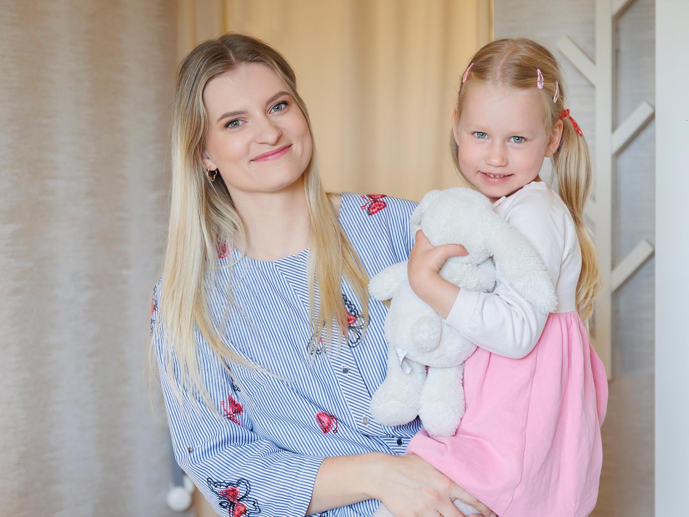A mother stands in a warmly lit room, smiling as they hold their daughter who is hugging a soft toy.