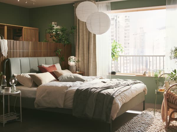 A muted green bedroom shows a bed with a two REGOLIT/HEMMA lamps hanging overhead. Outside the window are apartment blocks.