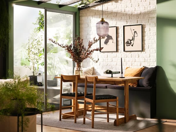 A NACKANÄS dining table and chairs is inside a room with a glass ceilng and doors. Sunlight streams onto plants in the room.
