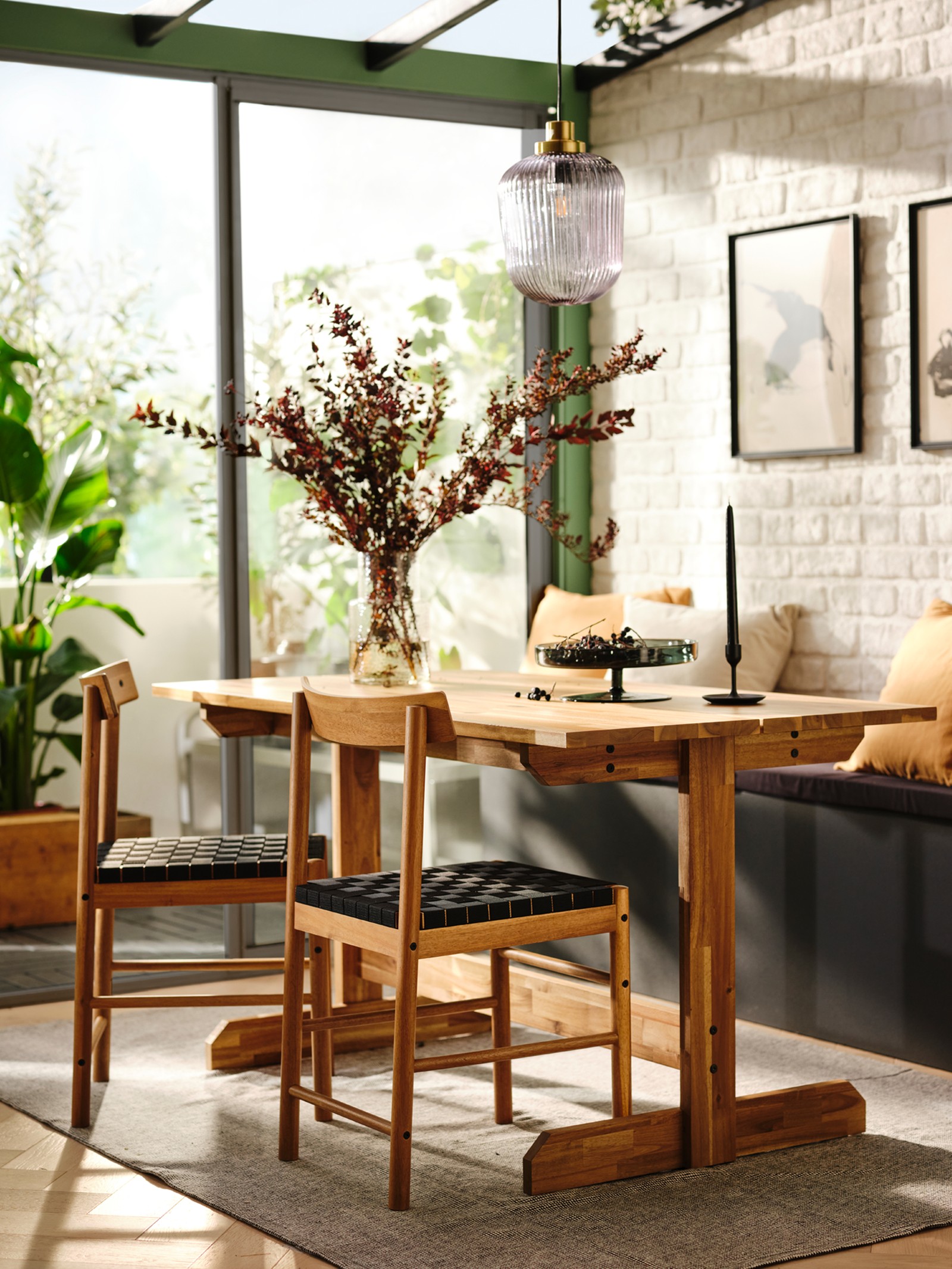A NACKANÄS table and two chairs in front of a bench with cushions on, and with a vase and a serving plate of fruit on top.