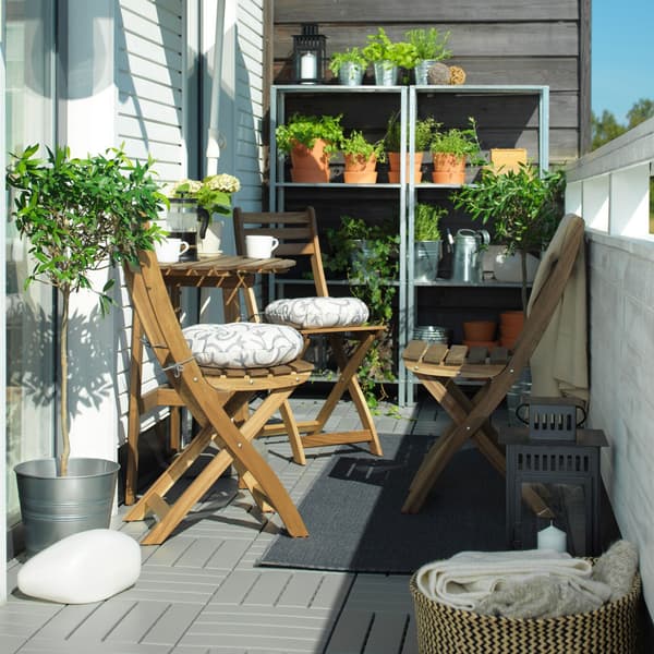 A narrow balcony with a wooden ASKHOLMEN table and chairs in the sun. Shelves with rows of plants can be seen behind.