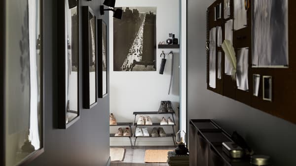 A narrow hallway with photos on the walls, black wired shoe storage stacked on top of each other, a black noticeboard.