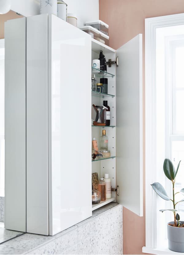 A narrow IKEA GODMORGON white wall cabinet, with glossy doors and 4 glass shelves stacked with bathroom supplies.