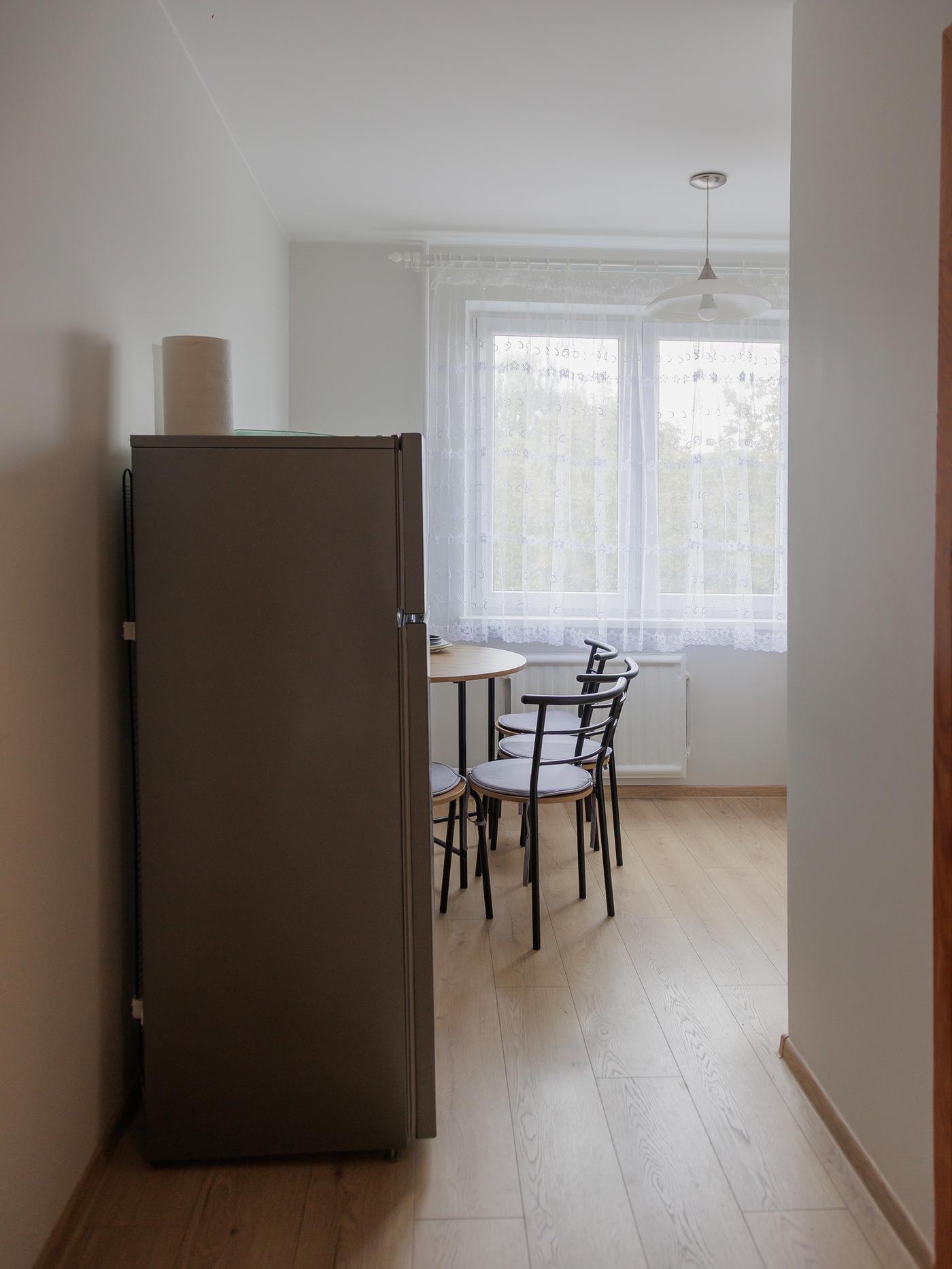 A narrow kitchen space with light wood flooring, a fridge and a small dining table near a window before the makeover.