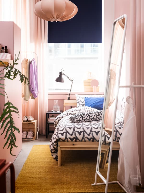 A narrow room in a rosy color scheme with a NEIDEN bed, a tall KNAPPER mirror and ample room to store and display clothes.