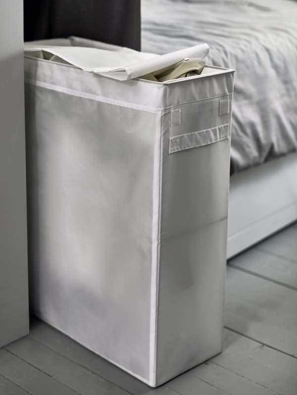 A narrow, white, laundry bag sits between a wall and the foot of a bed.
