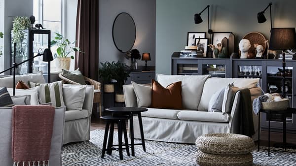 A natural BACKSÄLEN two-seat sofa with two black coffee tables in front of it and two grey HAUGA glass door cabinets behind.