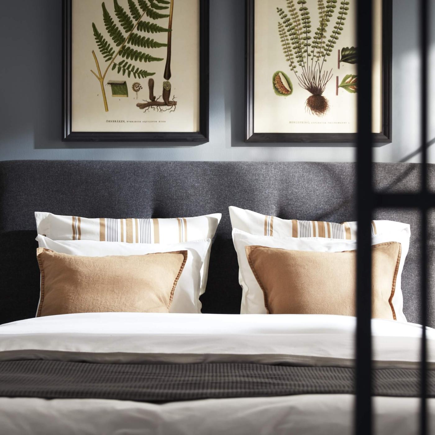 A neatly made bed with tan and striped pillows on it and plant pictures hanging above the upholstered bed frame.