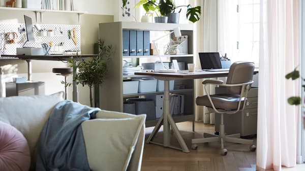 A neutral colour home workspace in the corner of a living room with two IDÅSEN sit/stand desks placed at different heights.