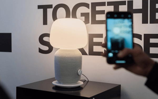 A new SYMFONISK speaker shaped like a table lamp sits on a stand and is photographed against a graphic sign.
